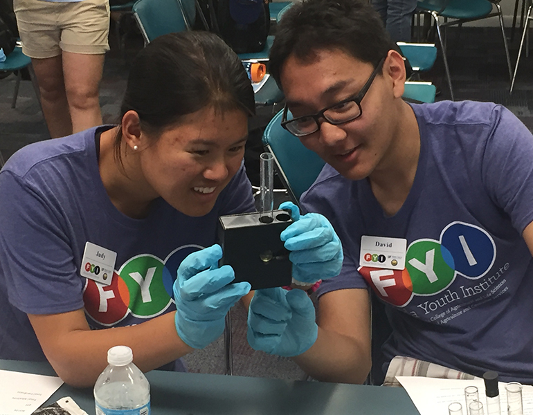 Two prospective CALS students wearing gloves and FYI shirts closely observe a test tube