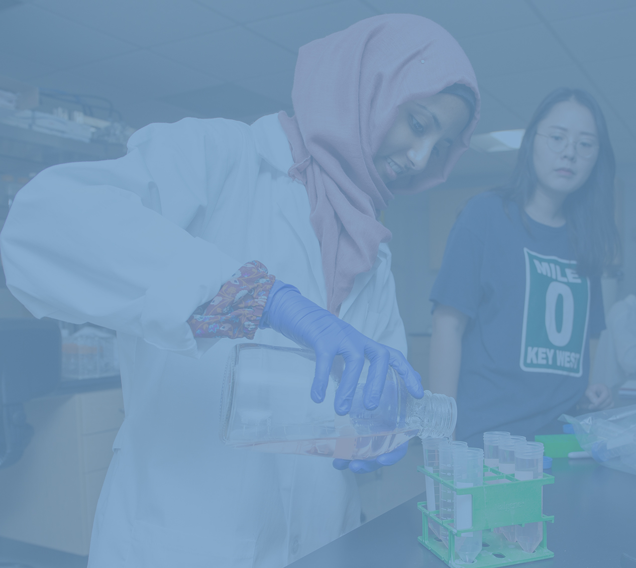 CALS student wearing a white lab coat, gloves and a headscarf pours a solution into graduated cylinders while her teacher observes.  